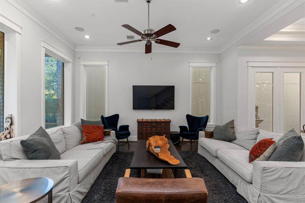 The spacious living room showcases large windows, recessed lighting, refined crown molding, a ceiling fan, and hardwood floors, all while offering a scenic view of the lush landscaping and mature trees.