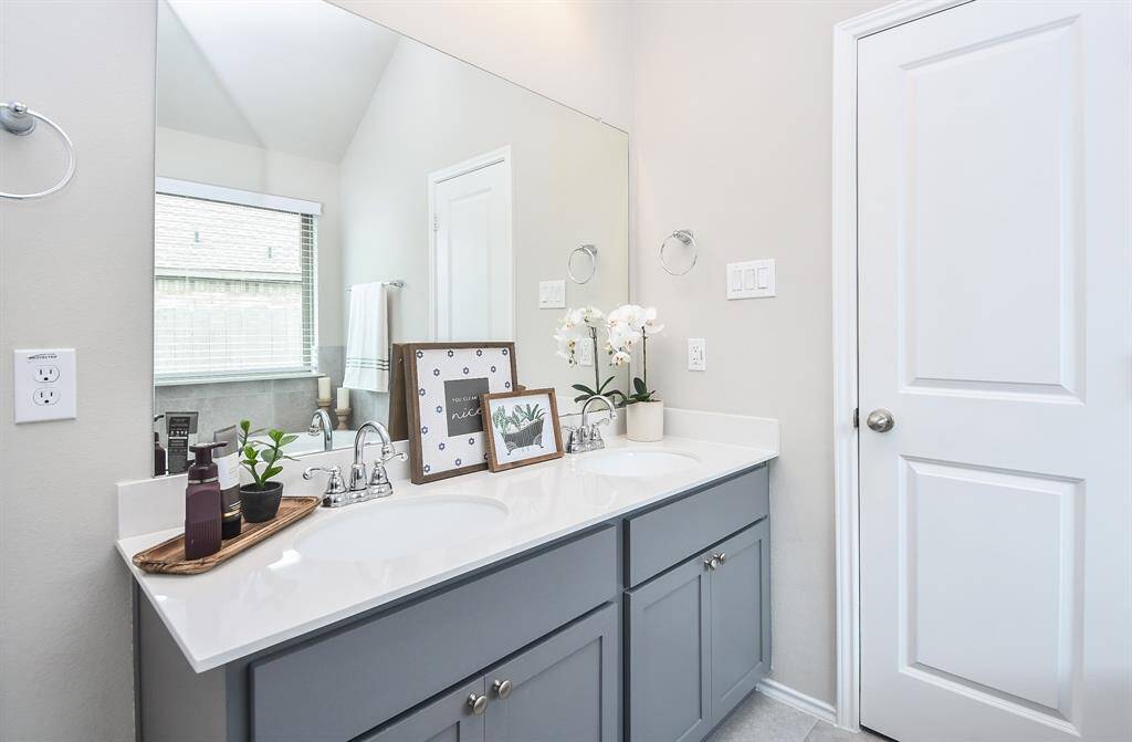 This bathroom features a modern dual-sink vanity with sleek gray cabinets and a large mirror. Natural light fills the space through a window, creating a bright and airy atmosphere. Elegant decor and neutral tones enhance its contemporary appeal.