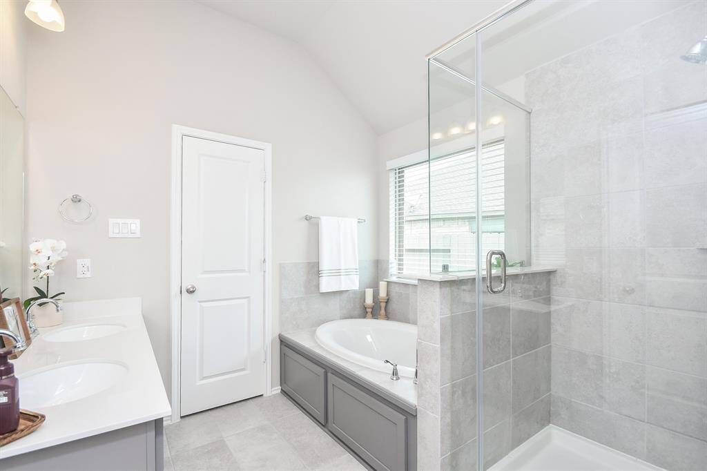 This bathroom also features a modern design with a glass-enclosed shower, a built-in bathtub, and a double vanity with ample counter space. The neutral tones and large window provide a bright and airy atmosphere.