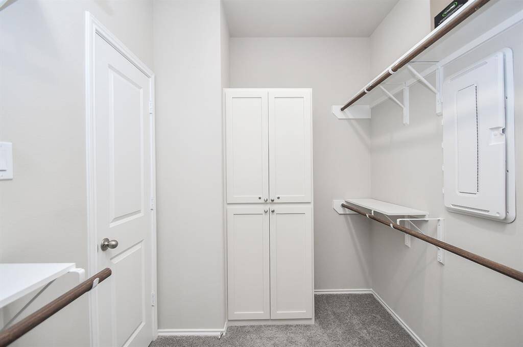 this primary spacious walk-in closet with white walls and carpeted flooring. It features built-in shelving, hanging rods, and a cabinet for extra storage, offering ample space for organization.