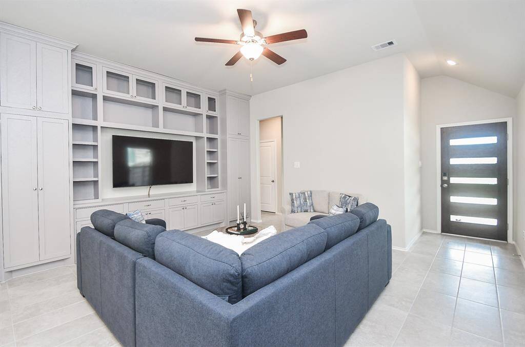 A spacious living room with a modern design featuring built-in shelving and cabinetry around a wall-mounted TV. It has a large, comfy sectional sofa, ceiling fan, and tile flooring. The entrance door is stylish and contemporary, allowing natural light to brighten the space.