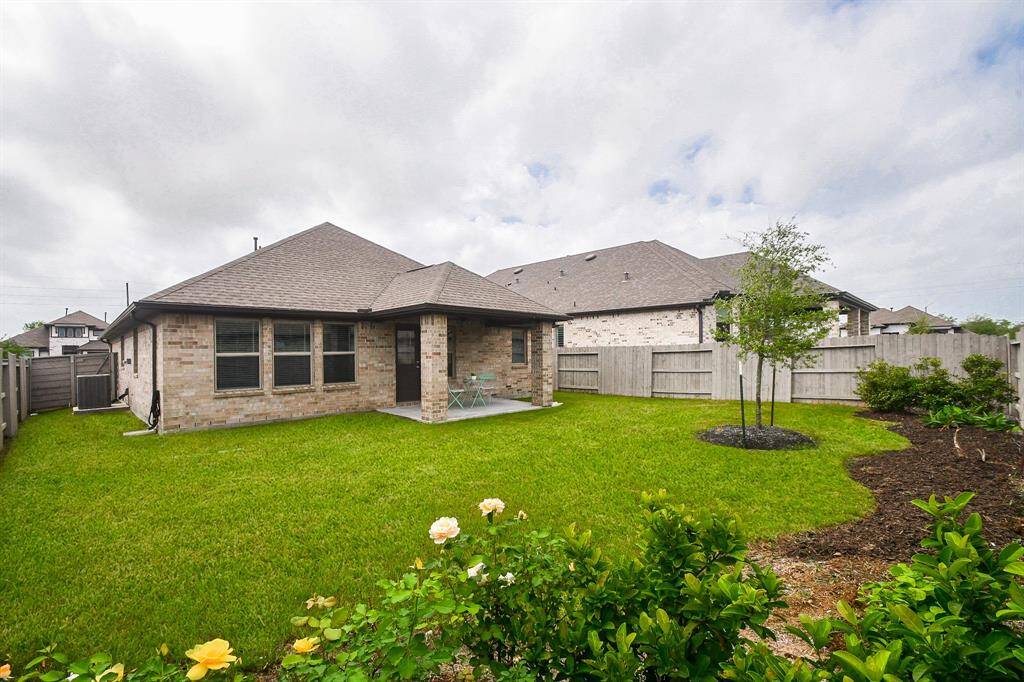 A charming backyard with a lush green lawn, bordered by a wooden fence for privacy. The brick house features a small covered patio, perfect for outdoor relaxation. Flowering plants and young trees add a touch of nature to the serene space.
