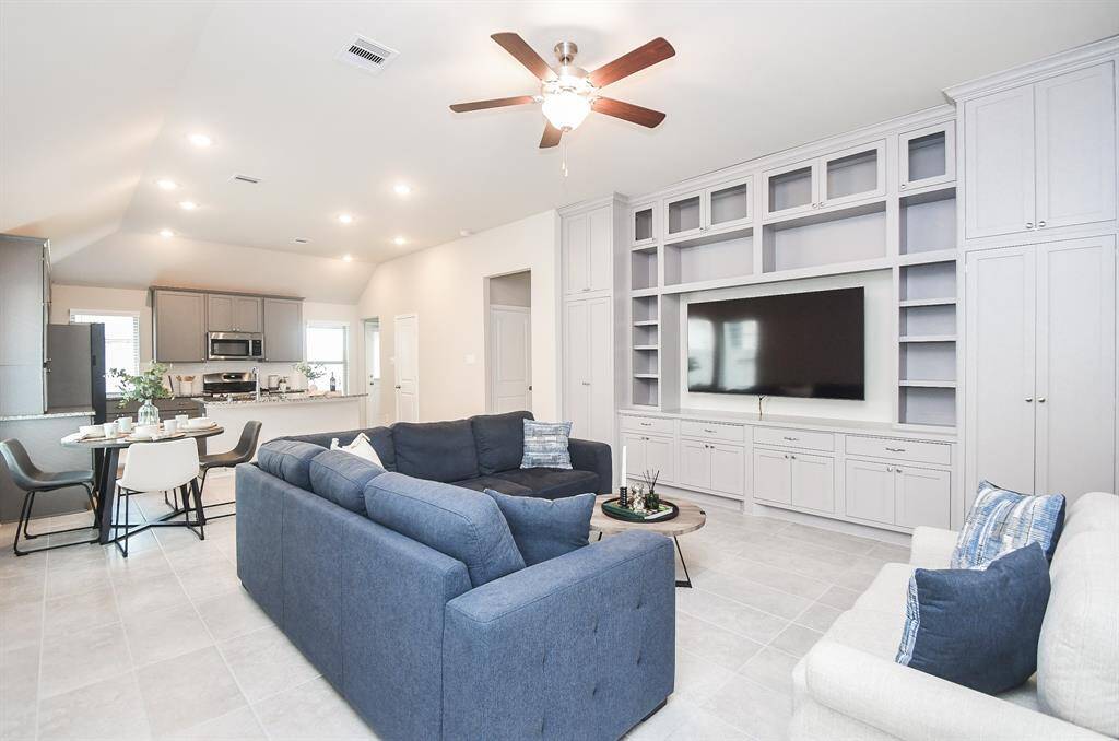 This open-concept living space features modern decor with a spacious blue sectional and built-in shelving surrounding a large TV. The adjacent kitchen has stainless steel appliances, ample cabinetry, and a cozy dining area, all under a ceiling fan and recessed lighting.
