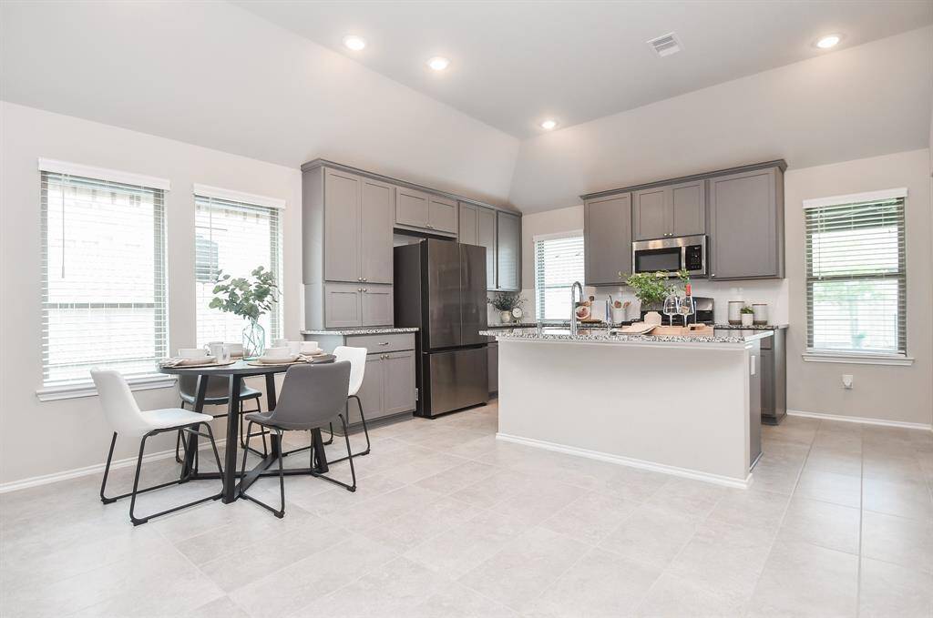 This modern kitchen features sleek gray cabinetry, stainless steel appliances, and a spacious island with bar seating. It has ample natural light from multiple windows and an adjacent dining area with a cozy table setting.