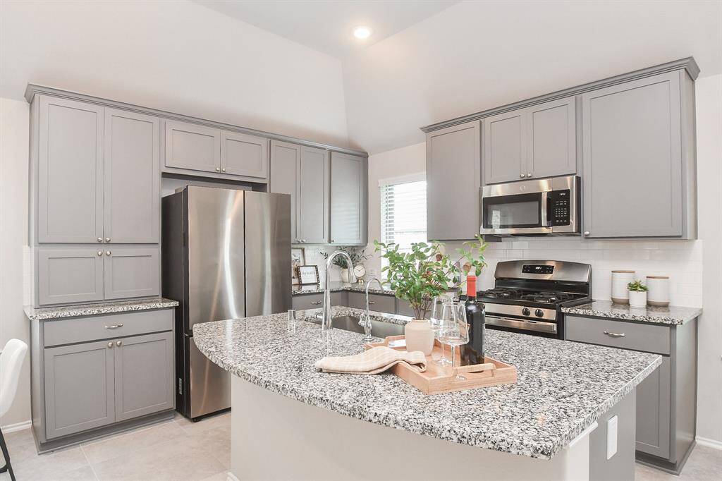 This modern kitchen features sleek gray cabinetry, granite countertops, and stainless steel appliances, including a double-door refrigerator and built-in microwave. An island with a sink provides additional workspace and seating, perfect for entertaining.