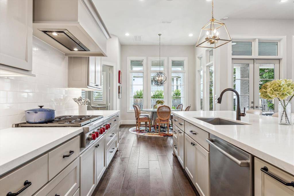This beautifully appointed kitchen blends refined design with everyday functionality, featuring sleek quartz countertops, custom cabinetry, and premium stainless steel appliances including a professional-grade gas range. The expansive island with farmhouse sink anchors the space, while wide-plank hardwood flooring and designer lighting add warmth and elegance. A wall of windows bathes the adjacent breakfast area in natural light, creating a bright and inviting atmosphere for both casual dining and entertain