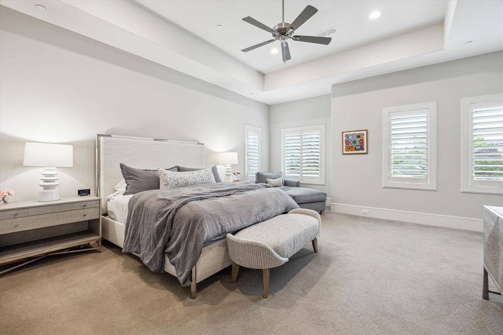 The serene primary suite offers a peaceful retreat with its soft palette, plush carpeting, and tray ceiling accentuating the spacious layout. A trio of shuttered windows invites natural light while maintaining privacy, creating a restful ambiance ideal for unwinding.