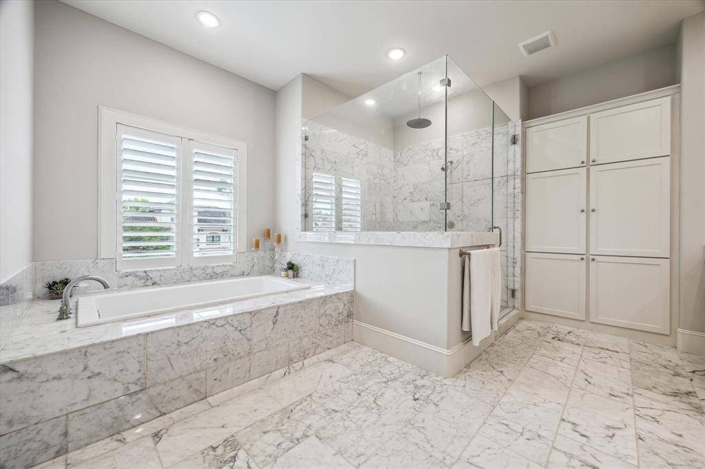 The spa-inspired primary bath exudes luxury with its floor-to-ceiling marble finishes, oversized frameless glass shower, and deep soaking tub perfectly positioned beneath shuttered windows for privacy and light. Generous built-in cabinetry adds refined function to this serene and elegant retreat.