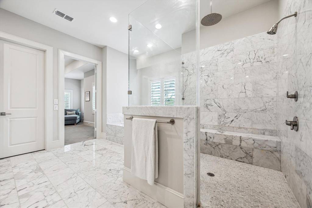 This luxurious primary bathroom showcases exquisite marble surfaces from floor to ceiling, offering a spa-like ambiance with a frameless glass walk-in shower featuring dual showerheads and a soothing rainfall fixture. A built-in marble bench and classic plantation shutters add comfort and privacy, while soft natural light enhances the serene, sophisticated aesthetic.