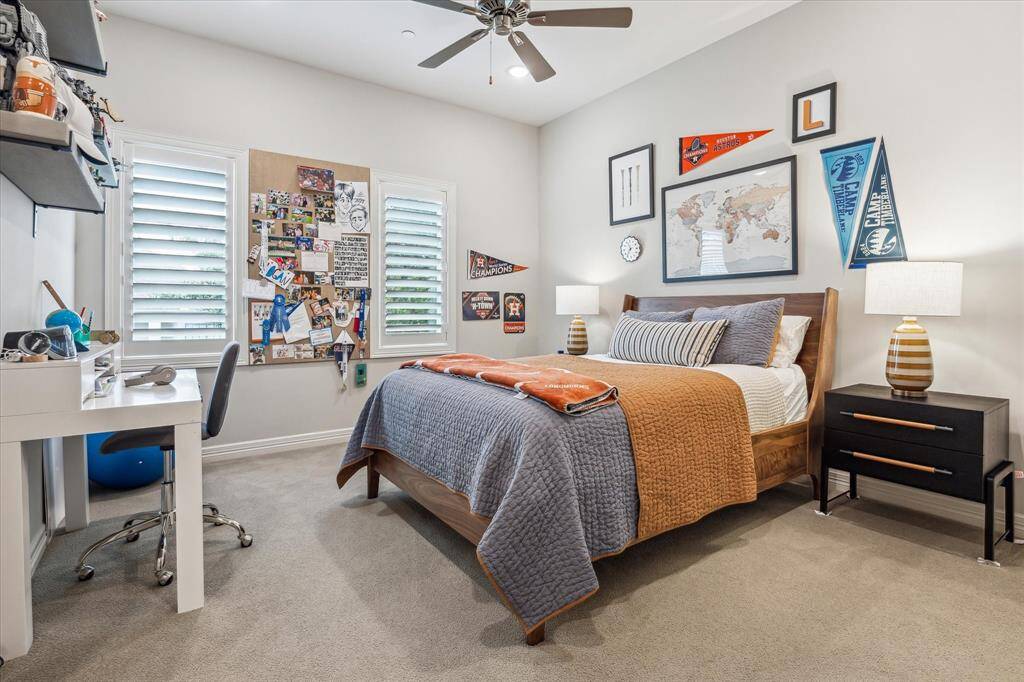 This inviting secondary bedroom offers a bright and cheerful atmosphere with plantation shutters, soft neutral walls, and plush carpeting underfoot. Thoughtfully designed with both comfort and function in mind, the space includes a dedicated desk area, ample natural light, and room for personal expression through custom wall decor.