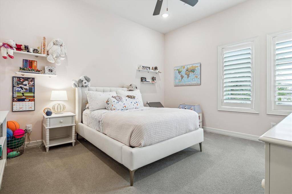 This bright and airy secondary bedroom features soft neutral tones, plush carpeting, and tall ceilings that enhance the sense of space, while dual shuttered windows provide abundant natural light and privacy.