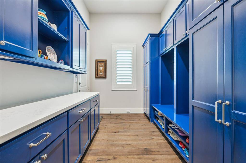 This vibrant mudroom combines bold design with everyday practicality, featuring striking custom blue cabinetry, ample built-in storage, and a durable quartz countertop—all set against warm hardwood floors and natural light from a shuttered window.