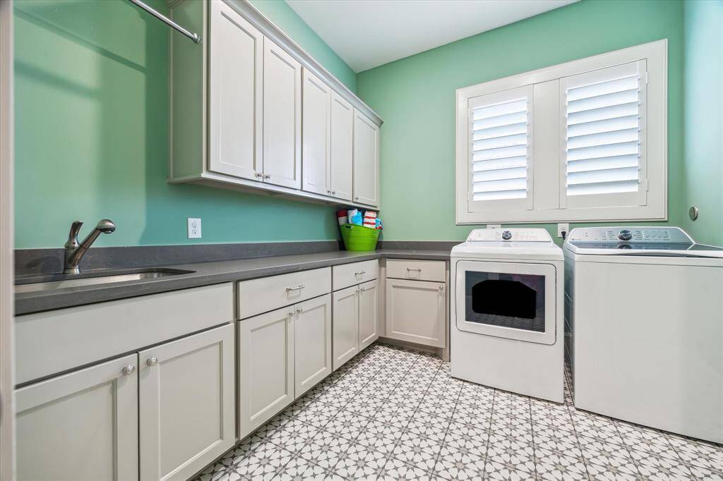 The laundry room combines charm and efficiency with patterned tile flooring, abundant cabinetry, quartz countertops, and a built-in sink—all illuminated by natural light through shuttered windows for a bright and cheerful workspace.
