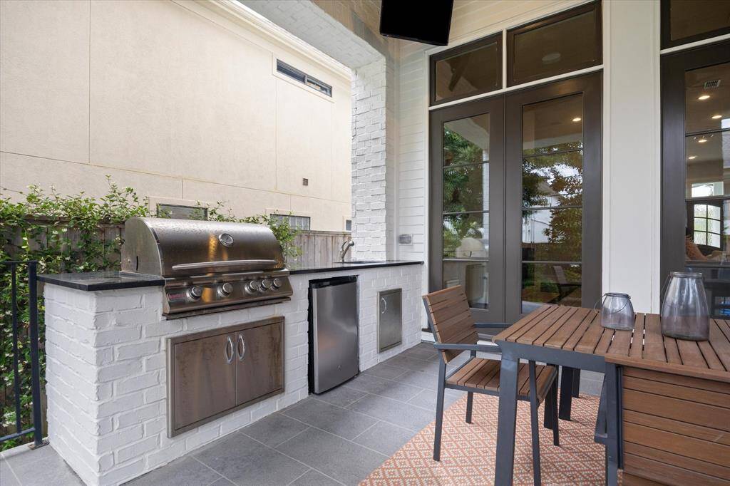 The outdoor kitchen is outfitted with a built-in stainless steel grill, under-counter refrigerator, and prep sink, all set against a backdrop of sleek granite countertops and painted brick—ideal for effortless entertaining and elevated alfresco dining.