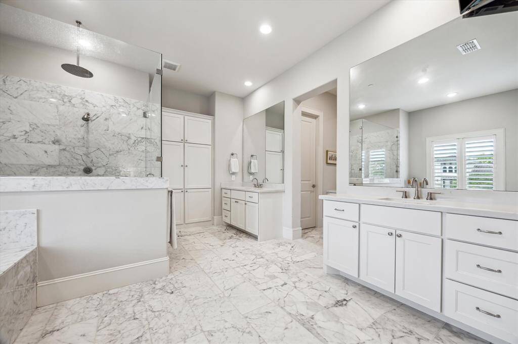 The primary bathroom exudes timeless sophistication with its polished marble flooring and shower surround, complemented by dual vanities and sleek brushed nickel fixtures. A frameless glass-enclosed rainfall shower and expansive mirrors enhance the sense of space and luxury, while crisp white cabinetry offers ample storage with a clean, elegant finish.