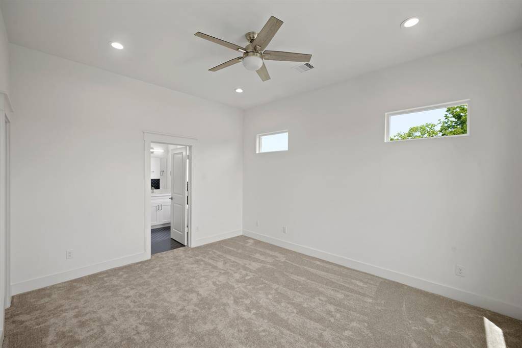 Bright, spacious bedroom featuring neutral carpeting, recessed lighting, and a ceiling fan. The room includes two small windows for natural light and an adjoining bathroom with modern fixtures.