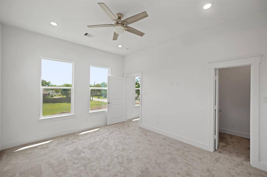 This bright bedroom features large windows, neutral carpet, a modern ceiling fan, and a small walk-in closet, creating a comfortable and airy space.