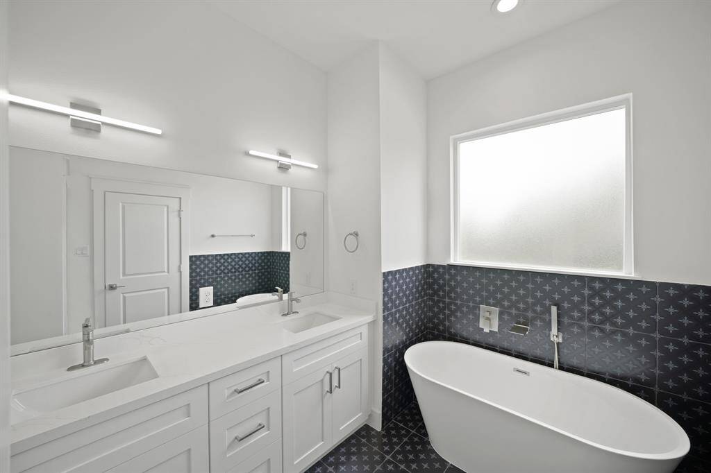 Bright and modern bathroom featuring a double vanity with sleek faucets, a freestanding bathtub, and stylish dark patterned tiles. The frosted window allows for natural light while maintaining privacy.