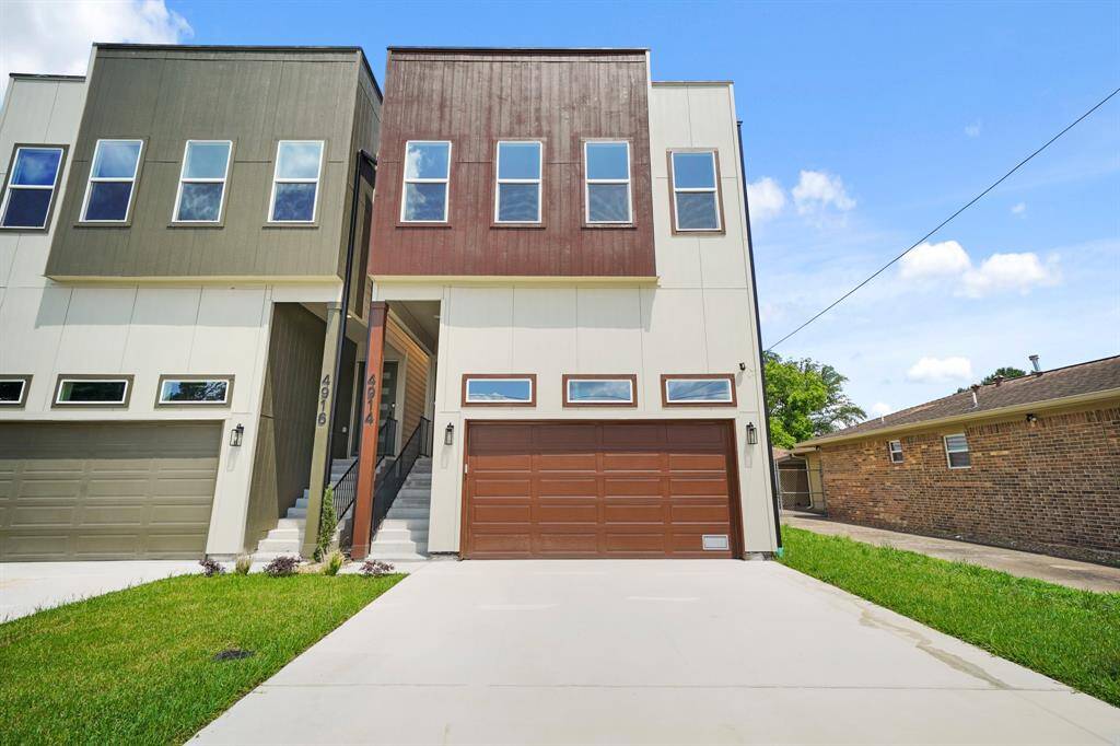 This modern home features a contemporary design with a two-tone exterior. It has a spacious garage, a small front yard, and a private driveway. Large windows offer ample natural light, and the entrance is elevated with a stylish staircase.