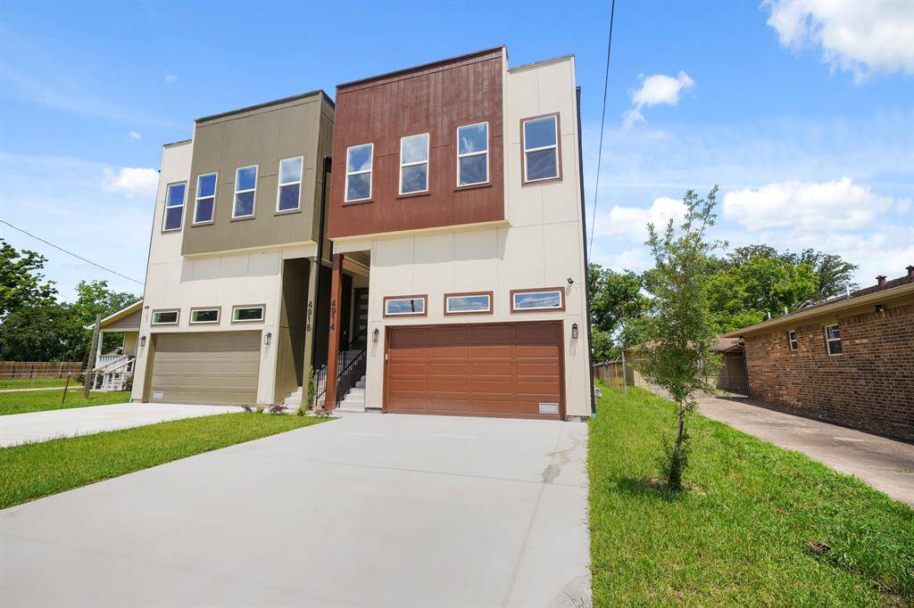 Modern Home with a sleek design, featuring two stories and a flat roof and two-car garage with large windows for natural light. The exterior color scheme is a mix of earth tones, and there's a spacious driveway with a small front lawn.