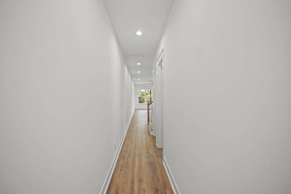 This photo shows a long, brightly lit hallway with white walls and wood flooring, leading to a window with a view of greenery. Recessed lighting adds a modern touch.