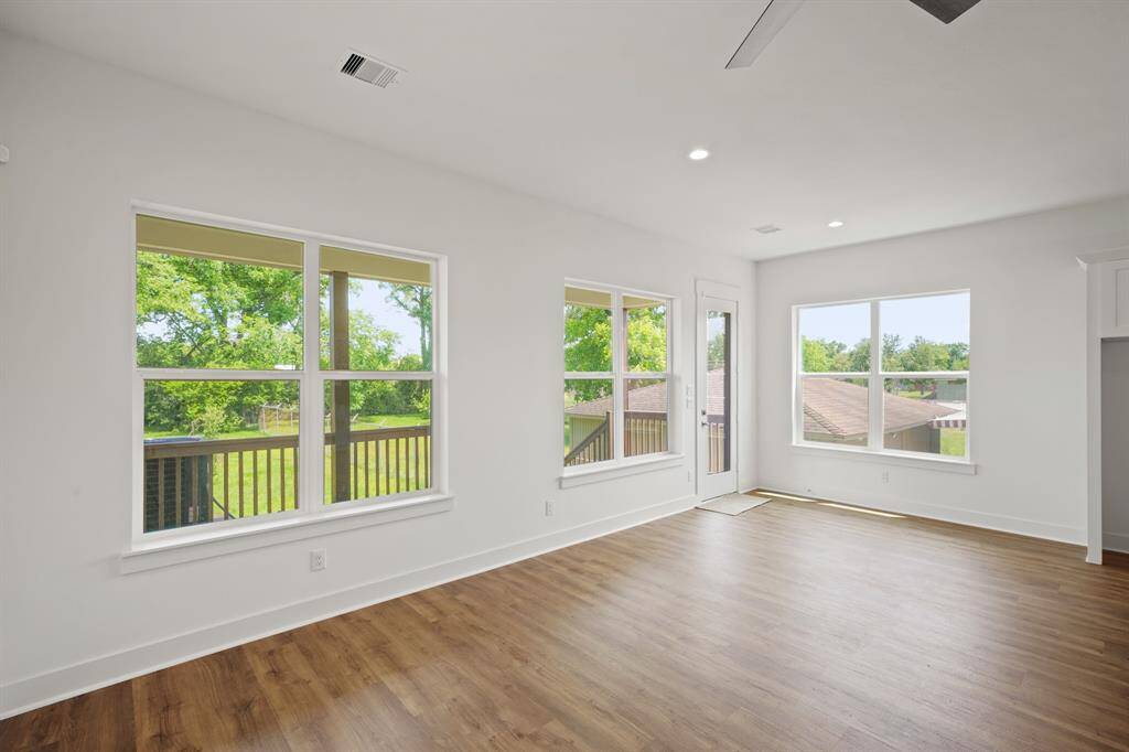 Bright and airy room with large windows offering lush green views. Features hardwood flooring, recessed lighting, and a ceiling fan, opening to a balcony for outdoor access. Ideal for a living space or sunroom.