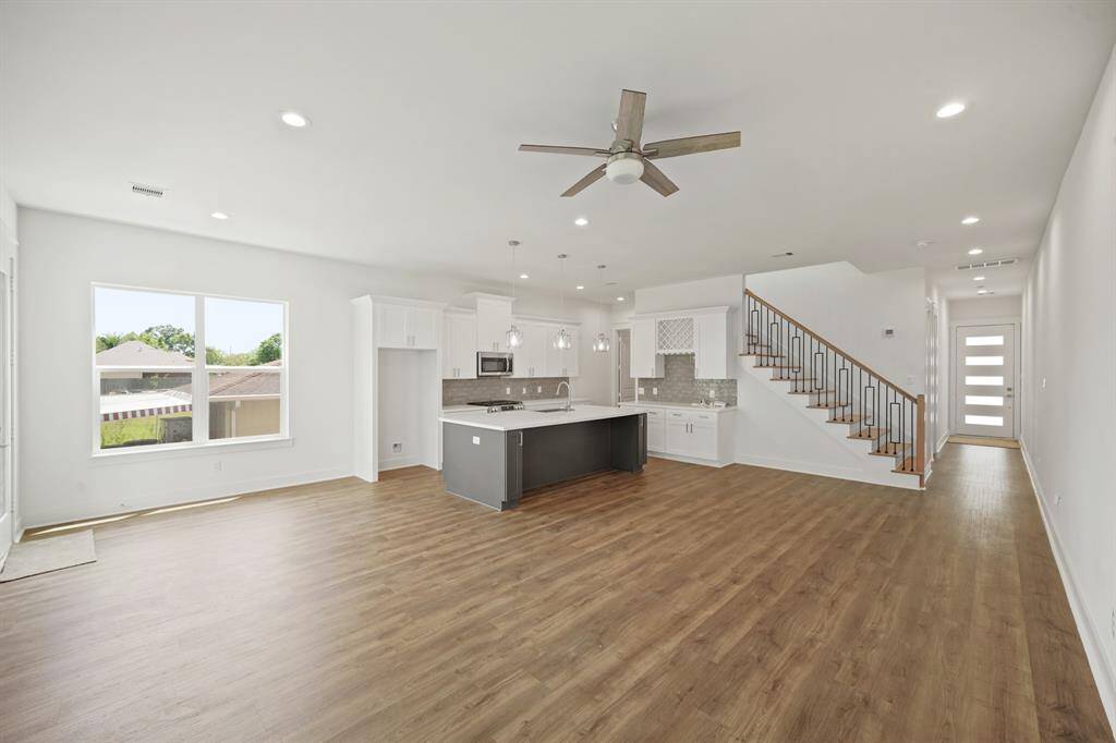 This open-concept living space features modern wood flooring, a sleek kitchen with white cabinetry and a central island, a ceiling fan, and ample natural light. A staircase leads to the upper level, and large windows offer a view of the backyard.