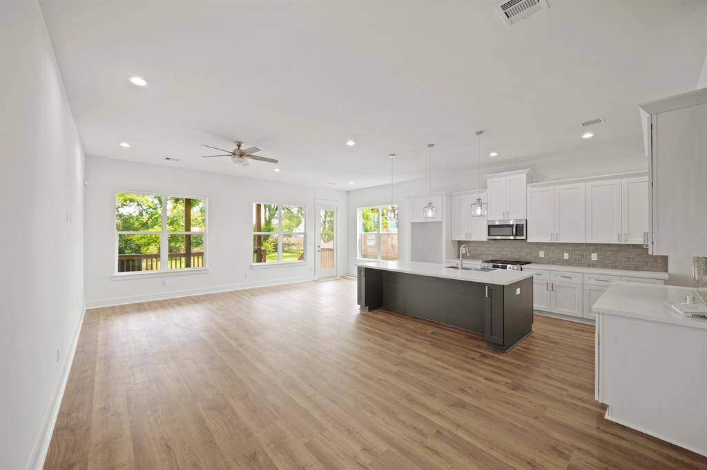 This photo showcases a spacious, open-concept living area with a modern kitchen featuring white cabinetry, a large island, and stainless steel appliances. The room has ample natural light from the large windows and wood flooring throughout, creating a bright and welcoming atmosphere.