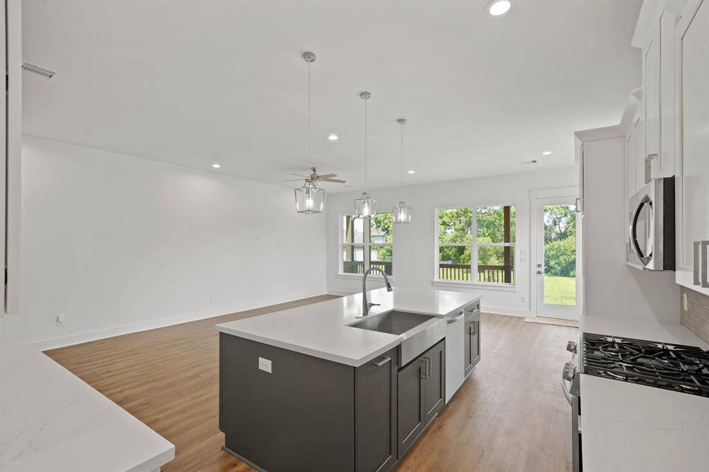 This open-concept kitchen and living area features sleek white cabinetry, a large central island with a sink, and modern light fixtures. The space is bright and airy, offering hardwood flooring and expansive windows that provide a scenic view of the greenery outside.