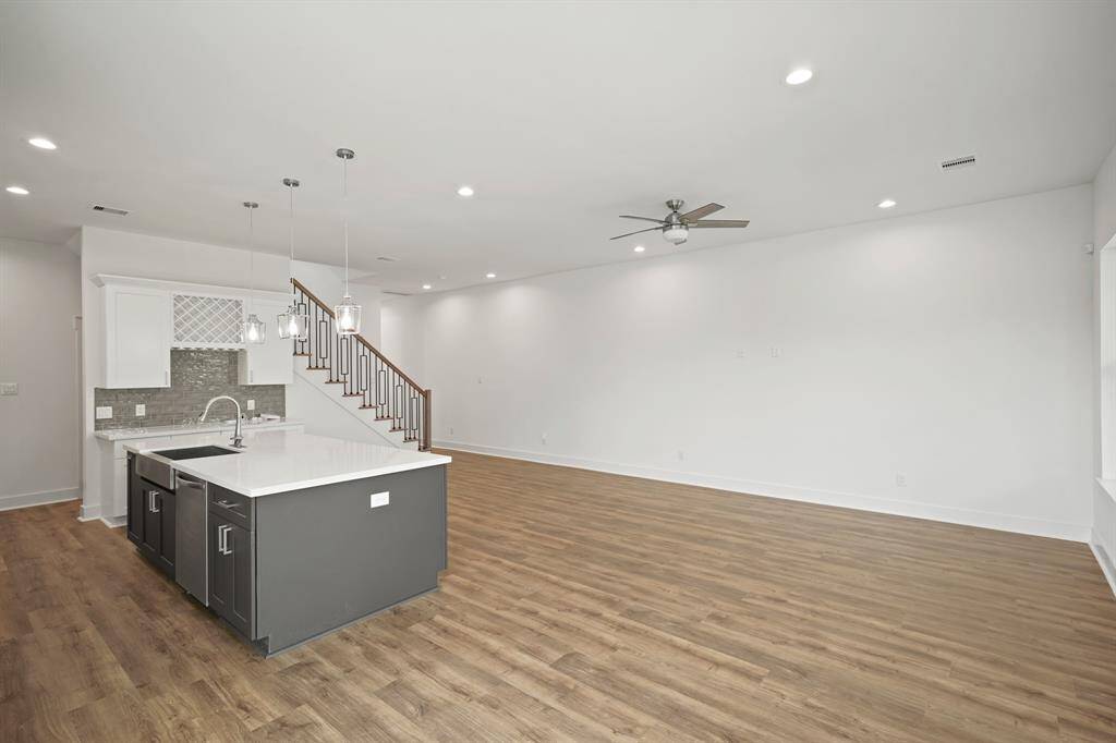 This photo showcases a spacious open-concept living area with modern hardwood floors, a sleek kitchen featuring a large island with pendant lighting, and a ceiling fan. The neutral color palette and recessed lighting create a bright, airy atmosphere, complemented by a staircase with a stylish railing.