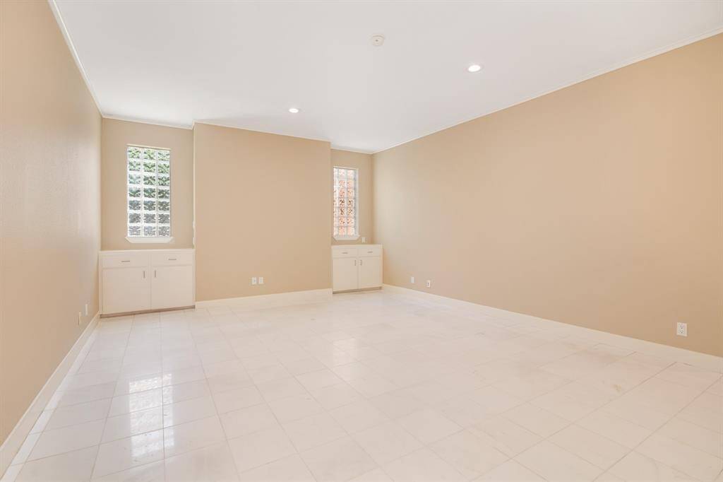 This is a spacious, well-lit room with beige walls and white tile flooring, featuring built-in white cabinetry under windows with a view of the exterior foliage.