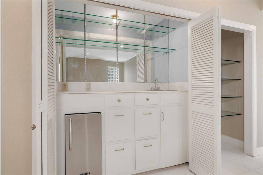 The living room features this compact wet bar area featuring white cabinetry, a sink, an under-counter refrigerator, and glass shelving. A mirrored backsplash and louvered closet doors add to the space's functionality and style.