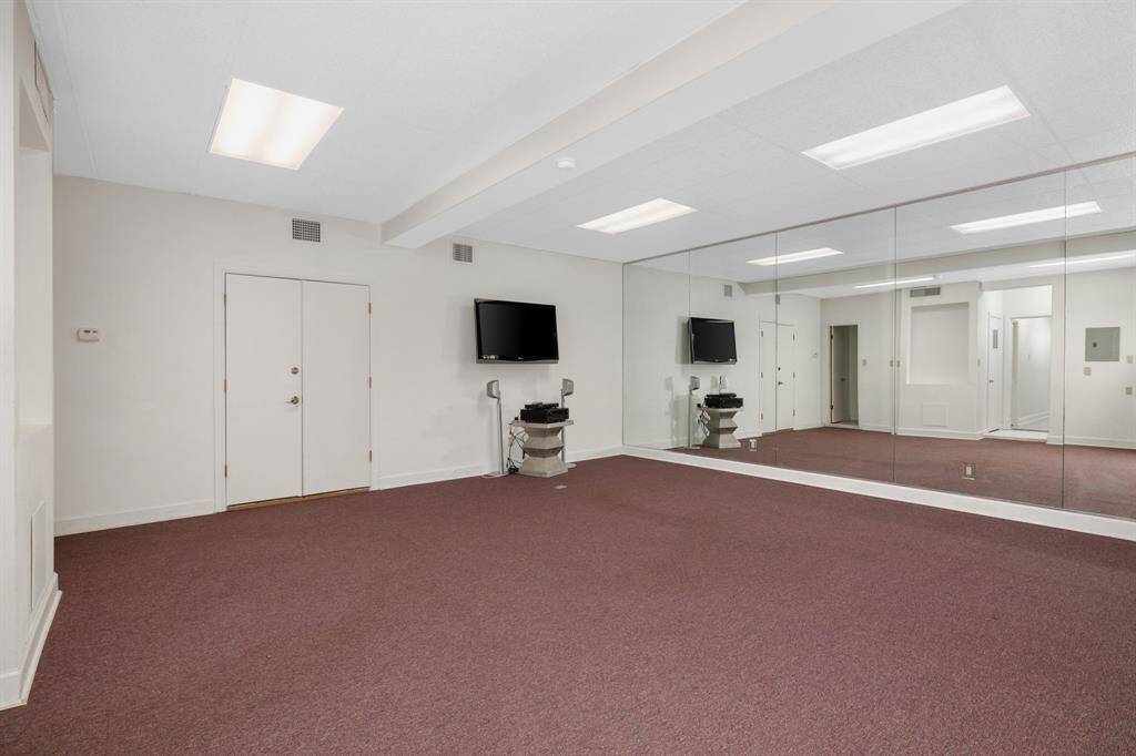 This is a spacious, well-lit room featuring mirrored walls, ideal for a home gym or dance studio, with plush carpeting and multiple doors for convenient access. It includes a mounted TV for entertainment or workout videos.