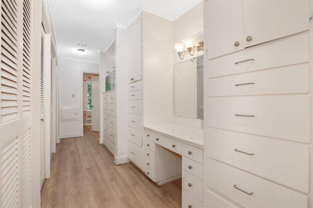 Walk in closet in primary suite. This is a well-lit, narrow space featuring multiple built-in white cabinets and drawers for ample storage, a vanity area with a large mirror, and wood-look flooring, leading to another room with a glimpse of a window.