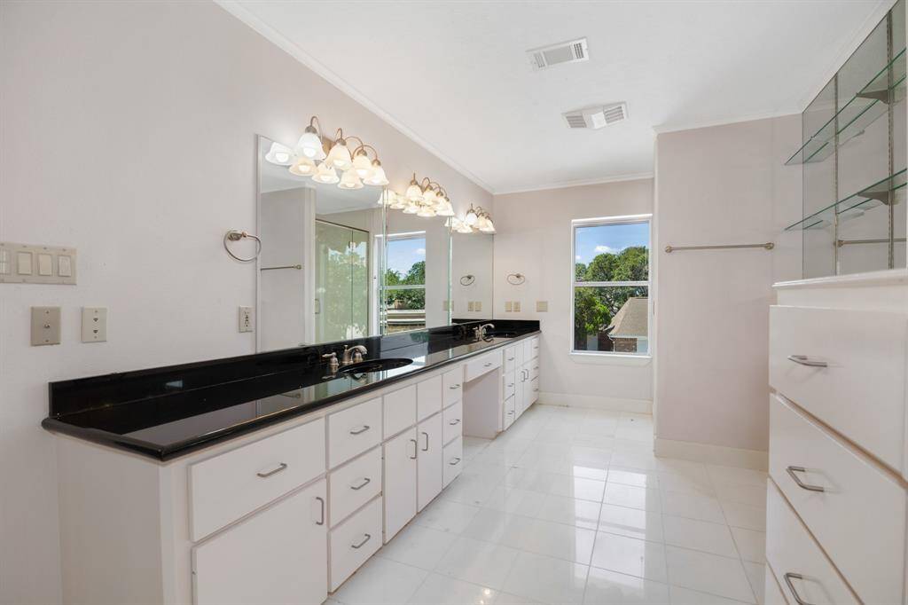 This is a bright, spacious bathroom featuring a long vanity with white cabinetry and black countertops, a large mirror with overhead lighting, a glass-enclosed shower, and a window allowing for natural light. In suite bath to large bedroom #2.