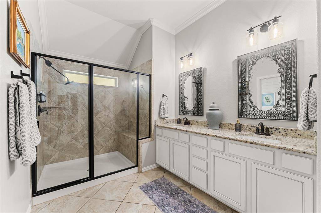 The Primary bathroom features a spacious walk-in shower with glass doors, a double vanity with granite countertops, stylish mirrors, and modern lighting. The neutral tones and tile flooring add a touch of elegance.
