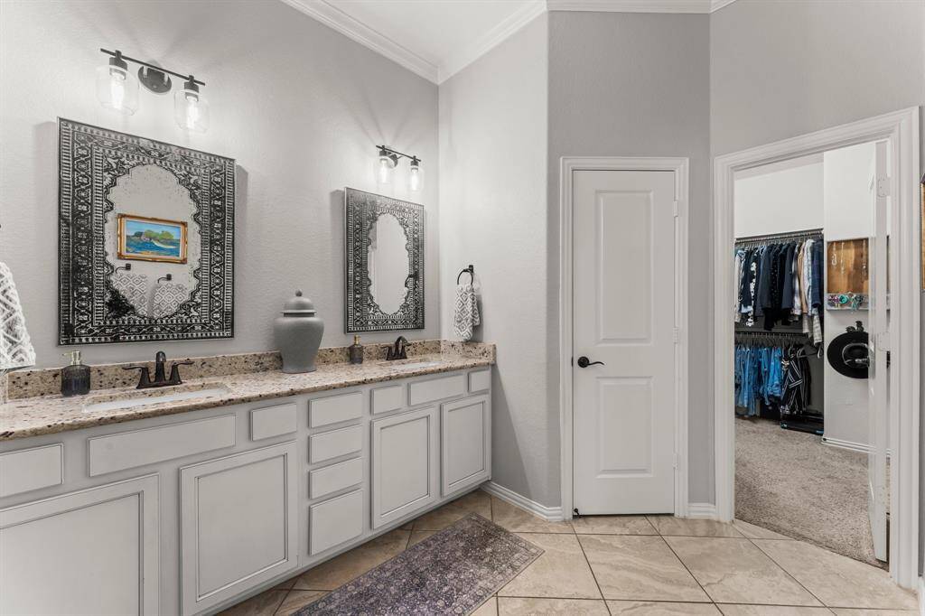 The primary bath offers  dual sinks, granite countertops, and decorative mirrors. The space features elegant lighting and neutral tile flooring. A door leads to a spacious walk-in closet, offering ample storage.