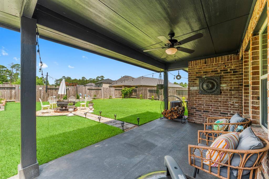 This inviting backyard features a covered patio with cozy seating, a ceiling fan, and a brick accent wall. The lush lawn leads to a charming fire pit area, perfect for outdoor gatherings.