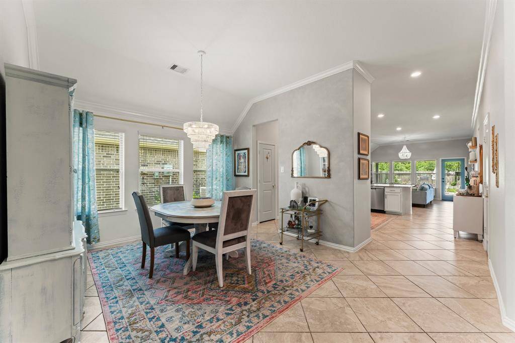 View from entry showcases a bright, open-concept dining and living area with tiled floors. The dining space features a round table under a modern chandelier and large windows with blue curtains. The living area extends into a kitchen with ample natural light and views of the backyard through multiple windows.