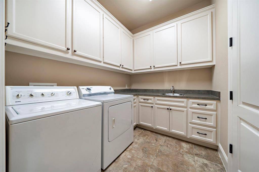 This photo shows a spacious laundry room featuring a washer and dryer, ample white cabinetry for storage, a countertop with a sink, and tile flooring.