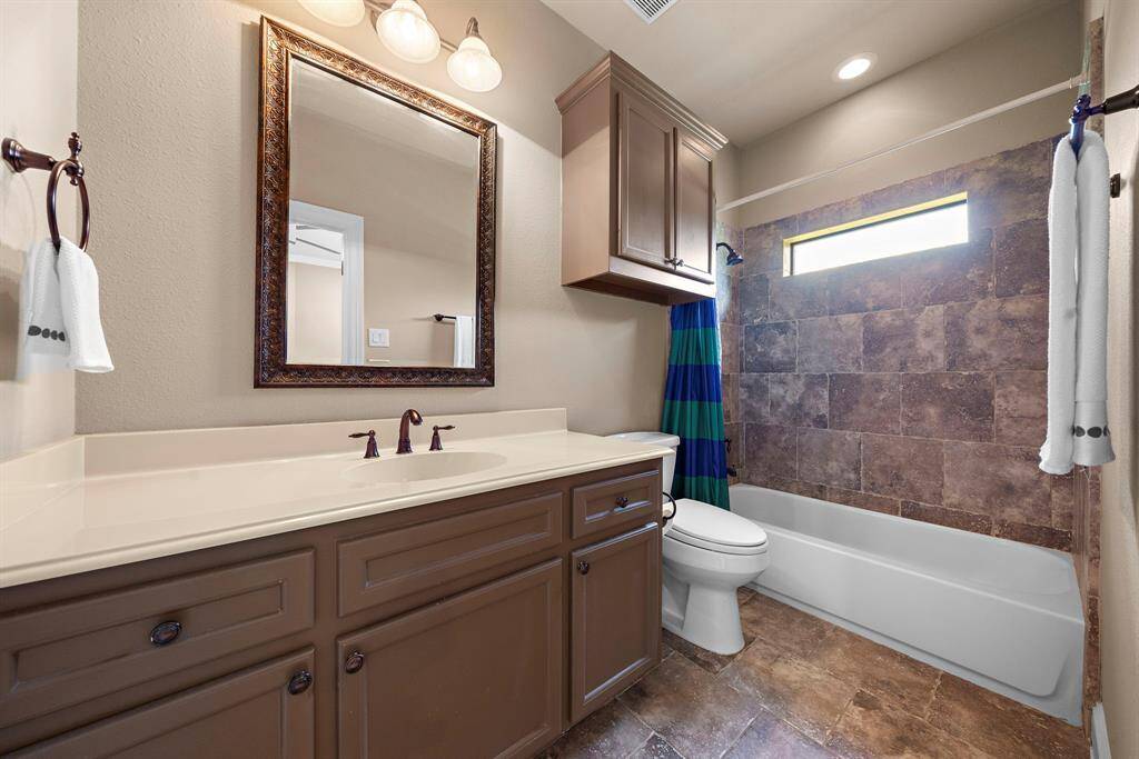This bathroom features a modern design with a large vanity, framed mirror, and bronze fixtures. It has a shower-tub combo with tiled walls, a small window for natural light, and ample storage with upper cabinets.