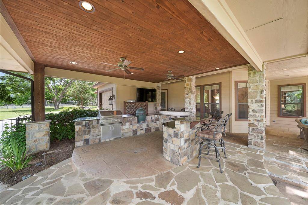 This outdoor patio features a covered area with a wooden ceiling and stone pillars, complete with a built-in grill and seating area. Ideal for entertaining, it overlooks a lush garden and offers a seamless indoor-outdoor living experience.