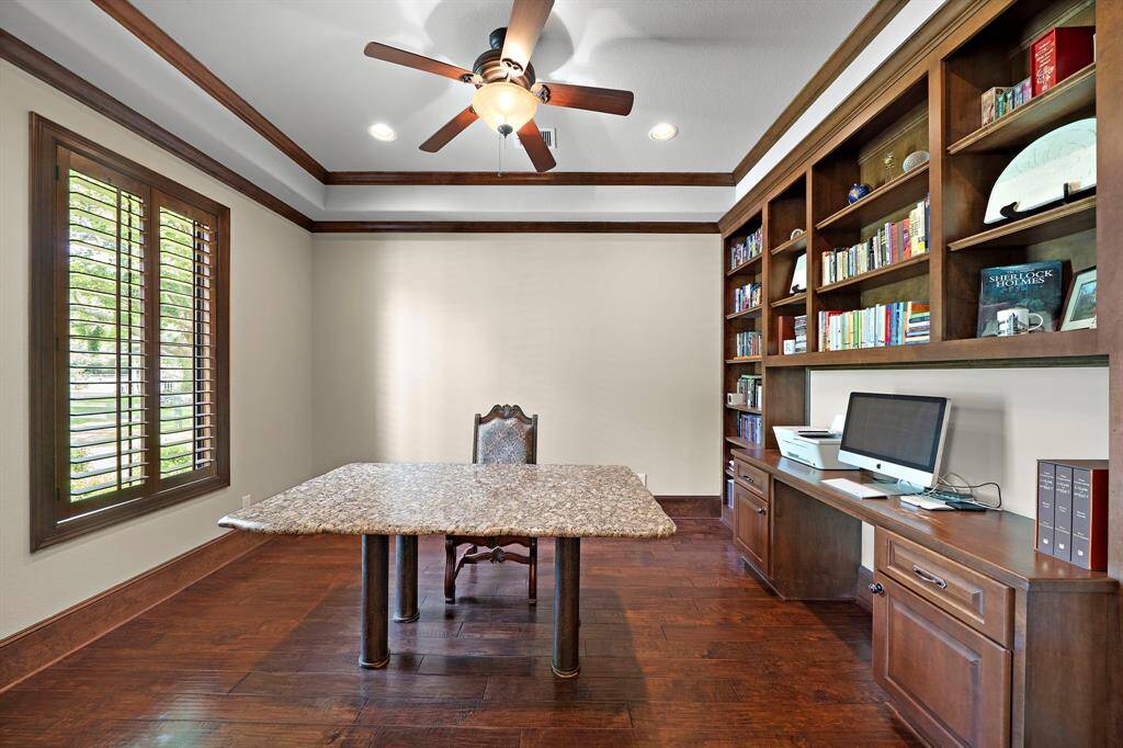 This home office includes elegant wooden built-in shelves and enough space for an oversized desk. The room is bright with natural light from a large window with wooden shutters. A ceiling fan adds comfort and style, complementing the rich wood floors and trim.