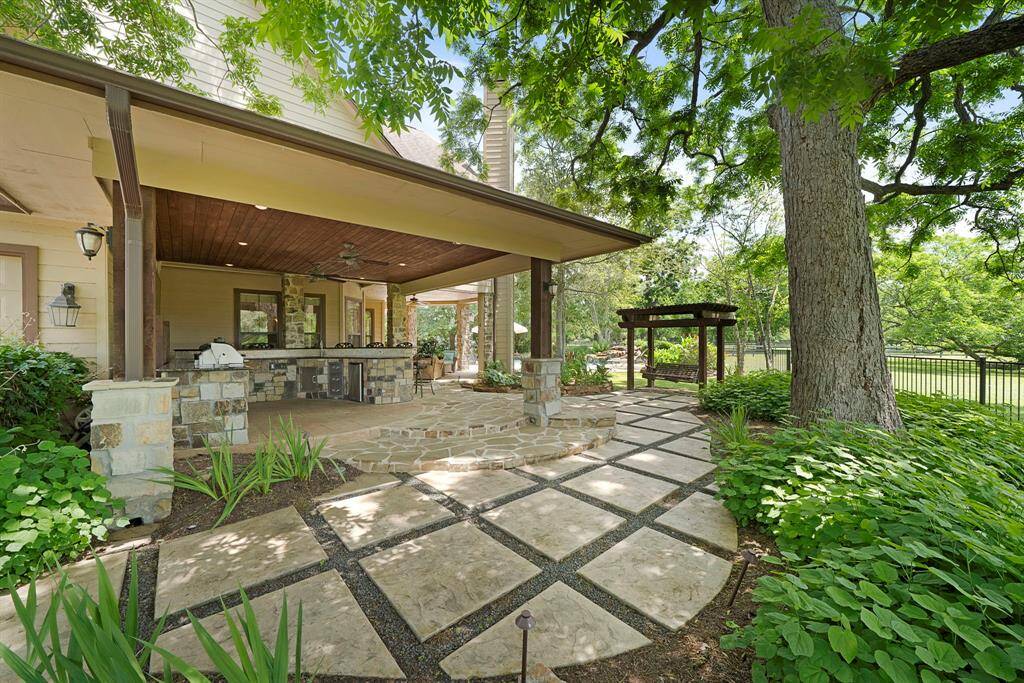 This photo showcases a charming outdoor living space with a covered patio featuring a built-in grill, sink, high top bar, and granite countertops. The area is surrounded by lush greenery, mature trees, a quaint swing, and a well-maintained garden, creating a serene and inviting atmosphere for relaxing or entertaining.