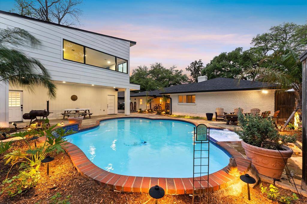 Welcome to the backyard oasis featuring a curvy swimming pool surrounded by a spacious stone patio. The area is accented with lush greenery, outdoor seating, and ambient lighting, offering a perfect setting for relaxation and entertaining.