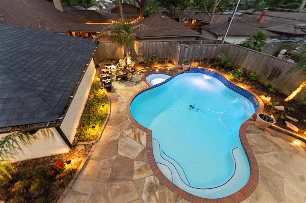 This inviting backyard features a uniquely shaped pool surrounded by a well-lit stone patio. There's a cozy seating area with a fire pit, perfect for entertaining. The space is enclosed by a wooden fence, providing privacy and a serene atmosphere with lush greenery and potted plants.