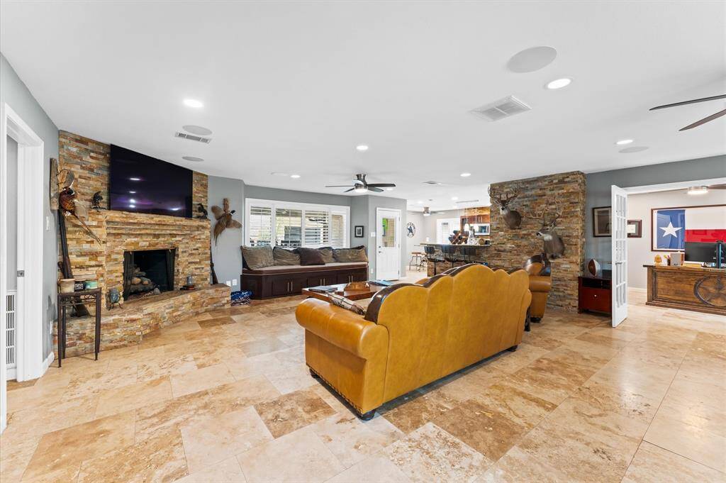 Ample living room with elegant stone fireplace, leather seating, and tiled flooring. Features mounted deer heads, large ceiling fans, and a wall-mounted TV. Ample natural light from multiple windows enhances the warm, inviting atmosphere.