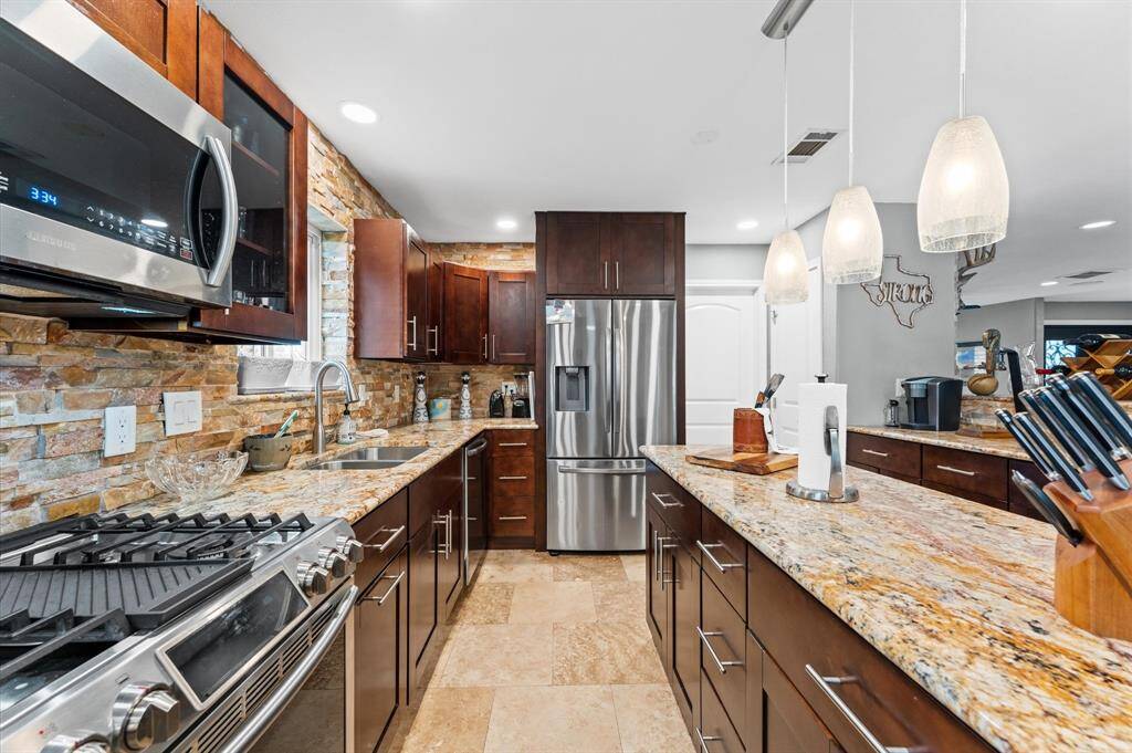 This photo showcases an open-concept kitchen and living area with modern design. The kitchen features dark wood cabinetry, stainless steel appliances, and a large island with granite countertops. The space flows into a living area with tile flooring, recessed lighting, and a stone fireplace.