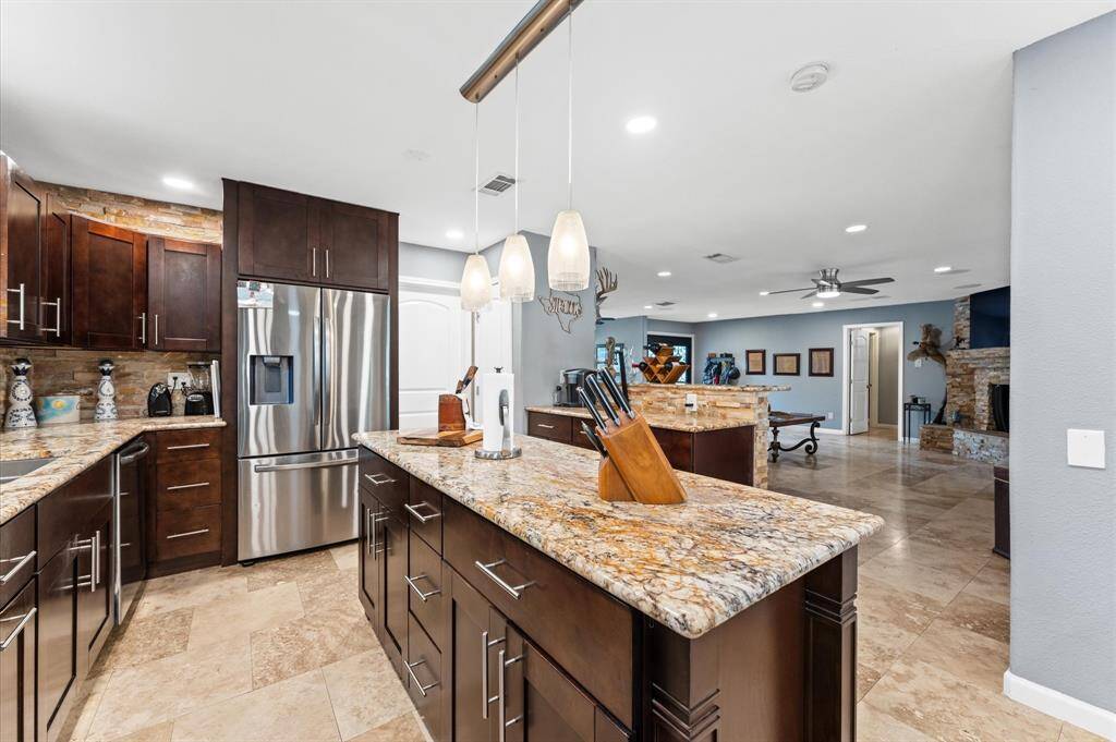 Elegant kitchen with granite countertops and dark wood cabinetry. It features a central island with pendant lighting, and breakfast bar stools. The open layout connects to a living area with a stone accent wall and ceiling fan, creating a spacious and inviting atmosphere.