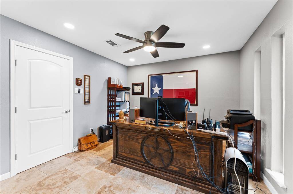 Cozy home office with a modern ceiling fan and ample natural light. Features a wooden desk, neutral walls, and tile flooring. Decor includes a large flag and shelving for storage.